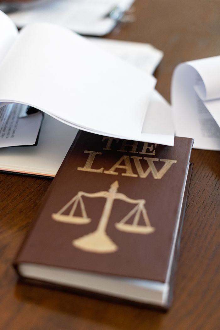 about-us-01 Close-up of a law book with scales icon on a wooden desk covered in papers.